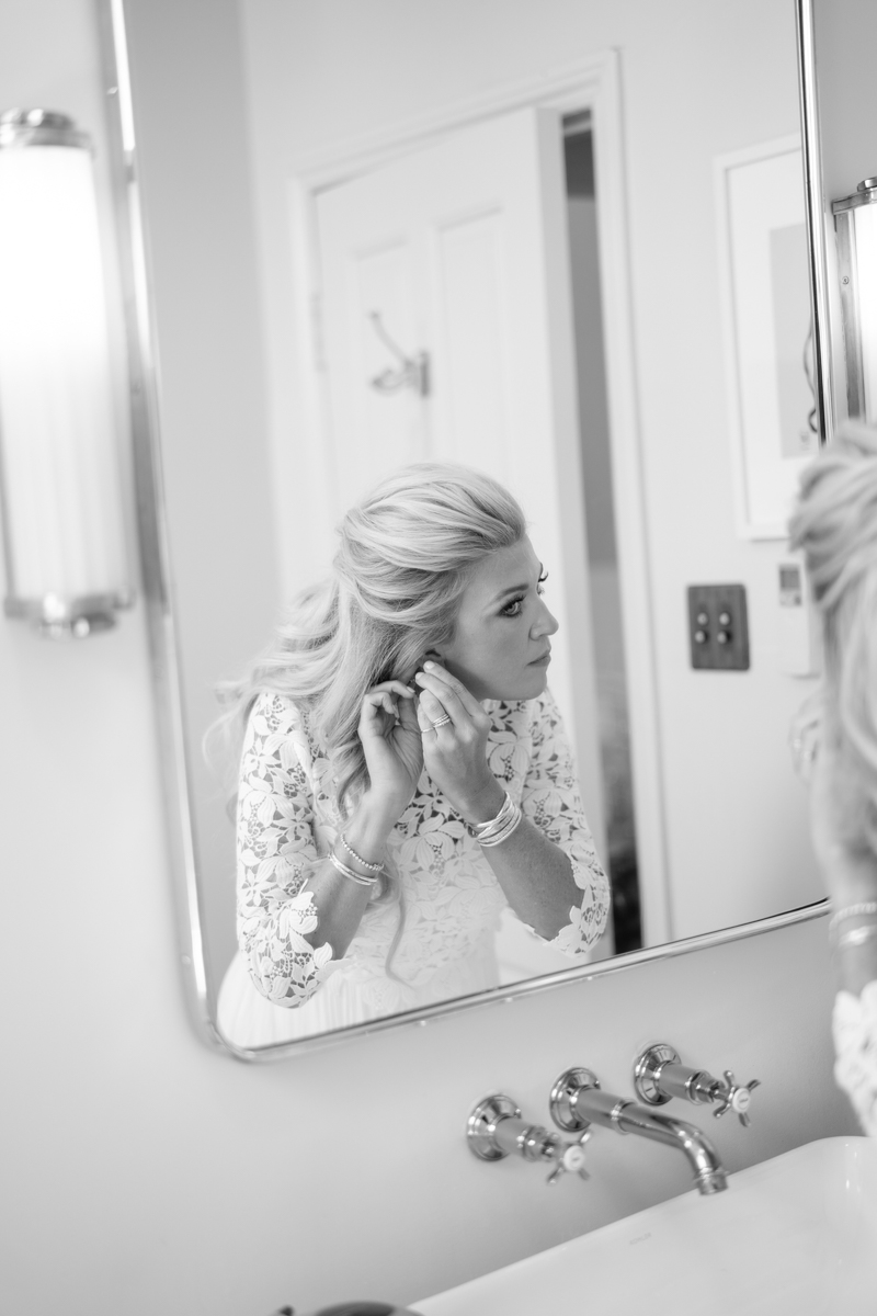 Black and white photo of bride looking in big mirror putting on pearls