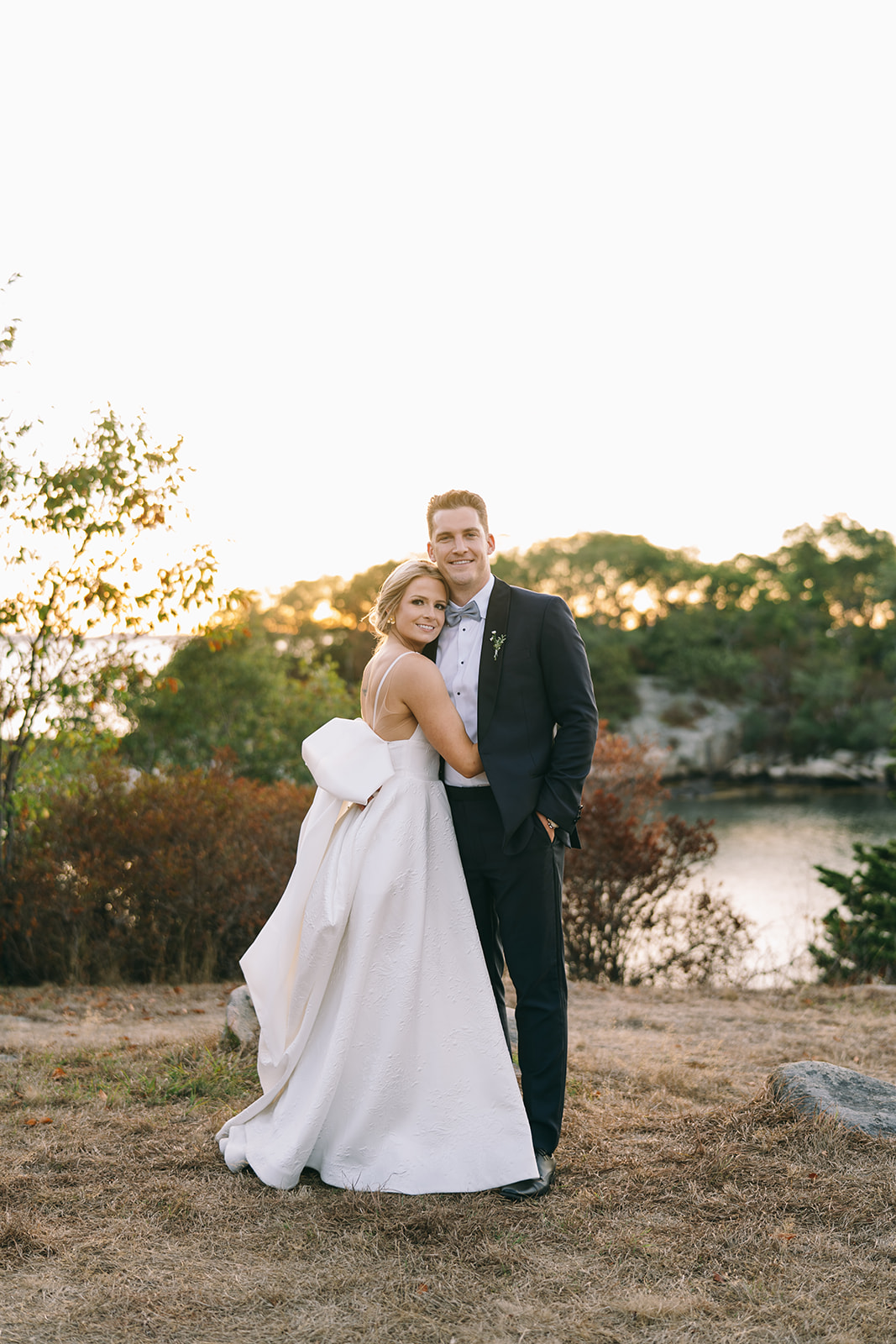 Bride with her arms around the groom with water in the background as the sun sets 