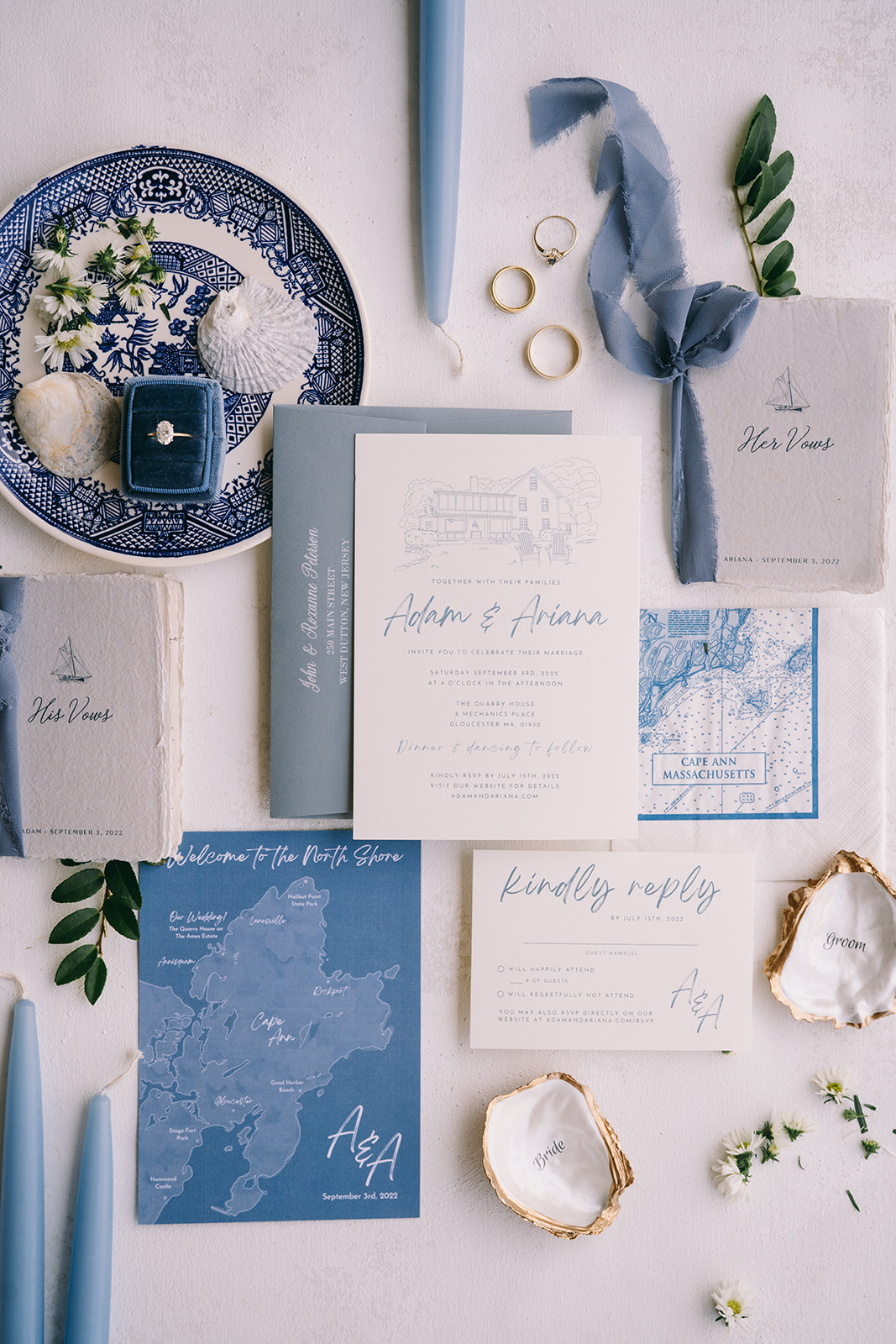 Wedding invitation flatlay with blue candles and gilded oyster shells near a blue china plate with flowers on it
