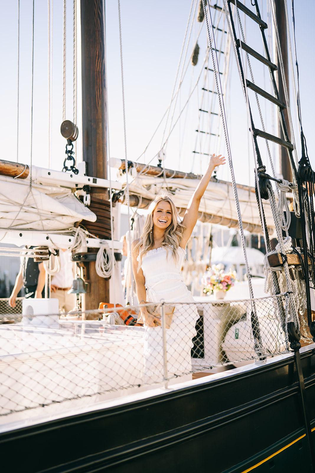 Woman standing on side of sailboat in white dress with her hand up in celebration