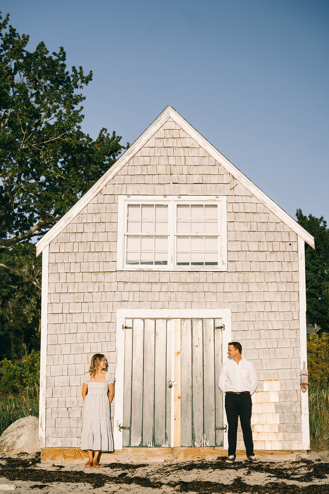 Man and woman standing in front of a small white barn on either side of the doors with a blue sky in the background