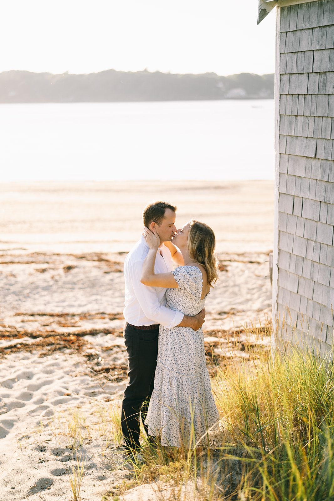 Woman in blue and white dress kissing a man in white shirt next to the barn with sand and ocean in the background