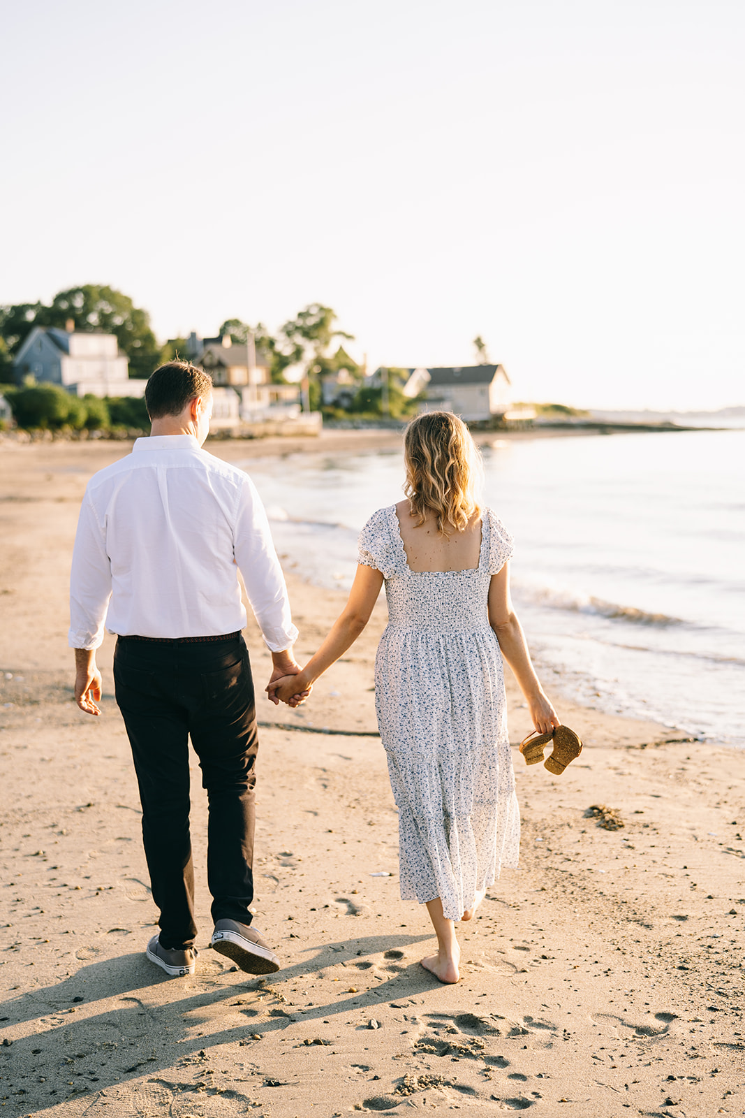 Couple walking along the coastline towards some houses and trees holding hands