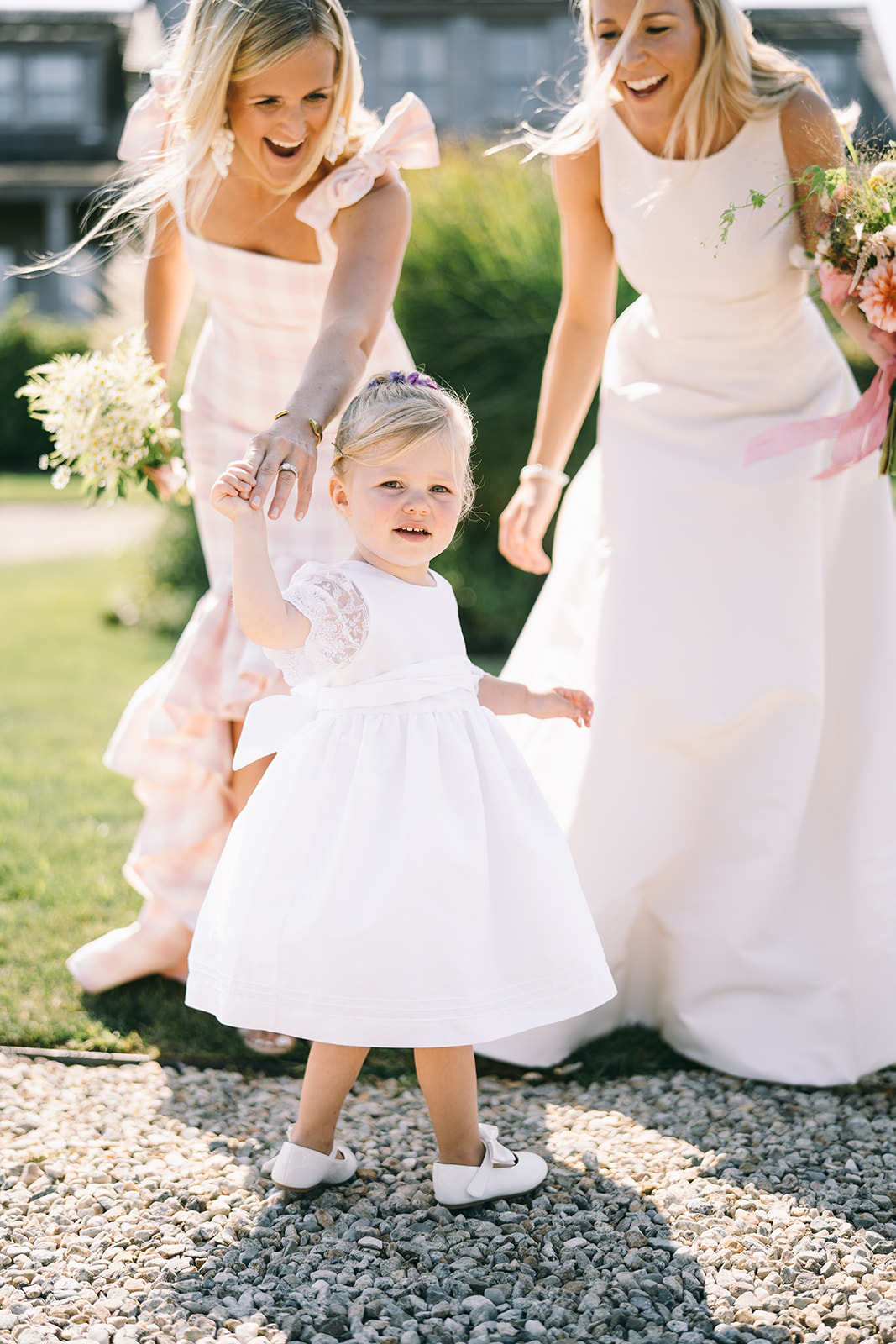 Woman in pink dress and woman in white dress leaning over and smiling at a small child in white