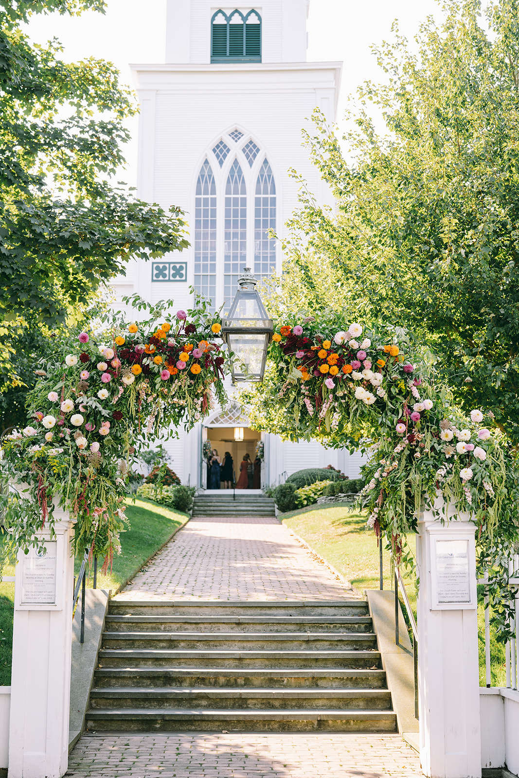 Outside of a church in Nantucket with a flower arch and green trees on either side of the walkway