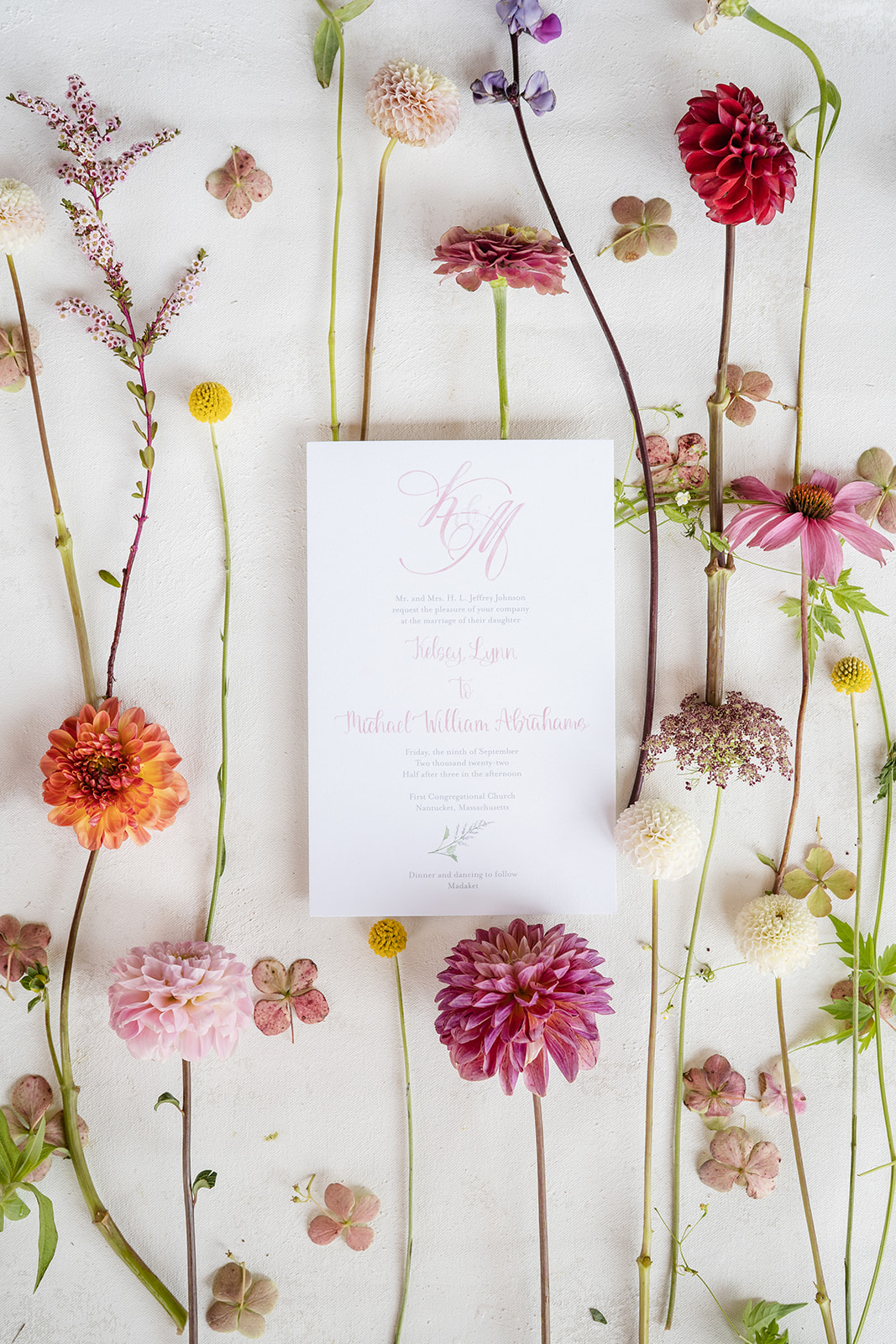 Wedding invitation on white paper with pink font flat layed on flowers with long, delicate stems