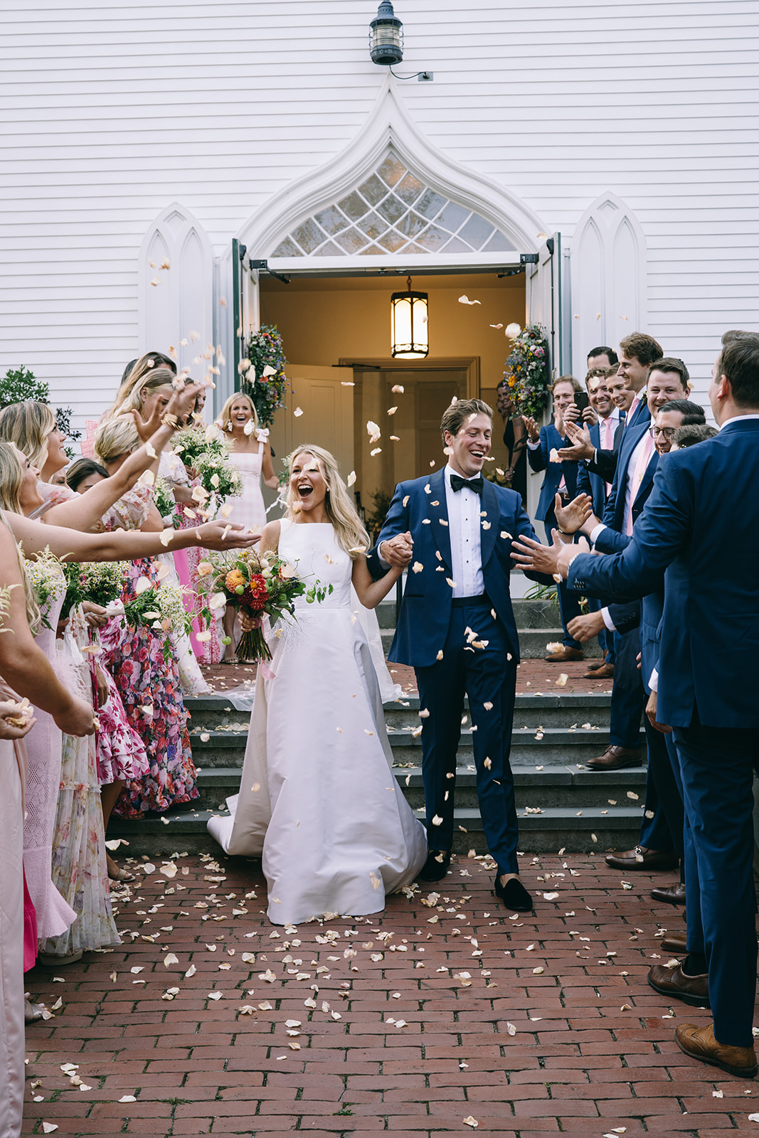 Woman in white dress and man in blue suit leaving church as people in colorful outfits shower them with flower petals