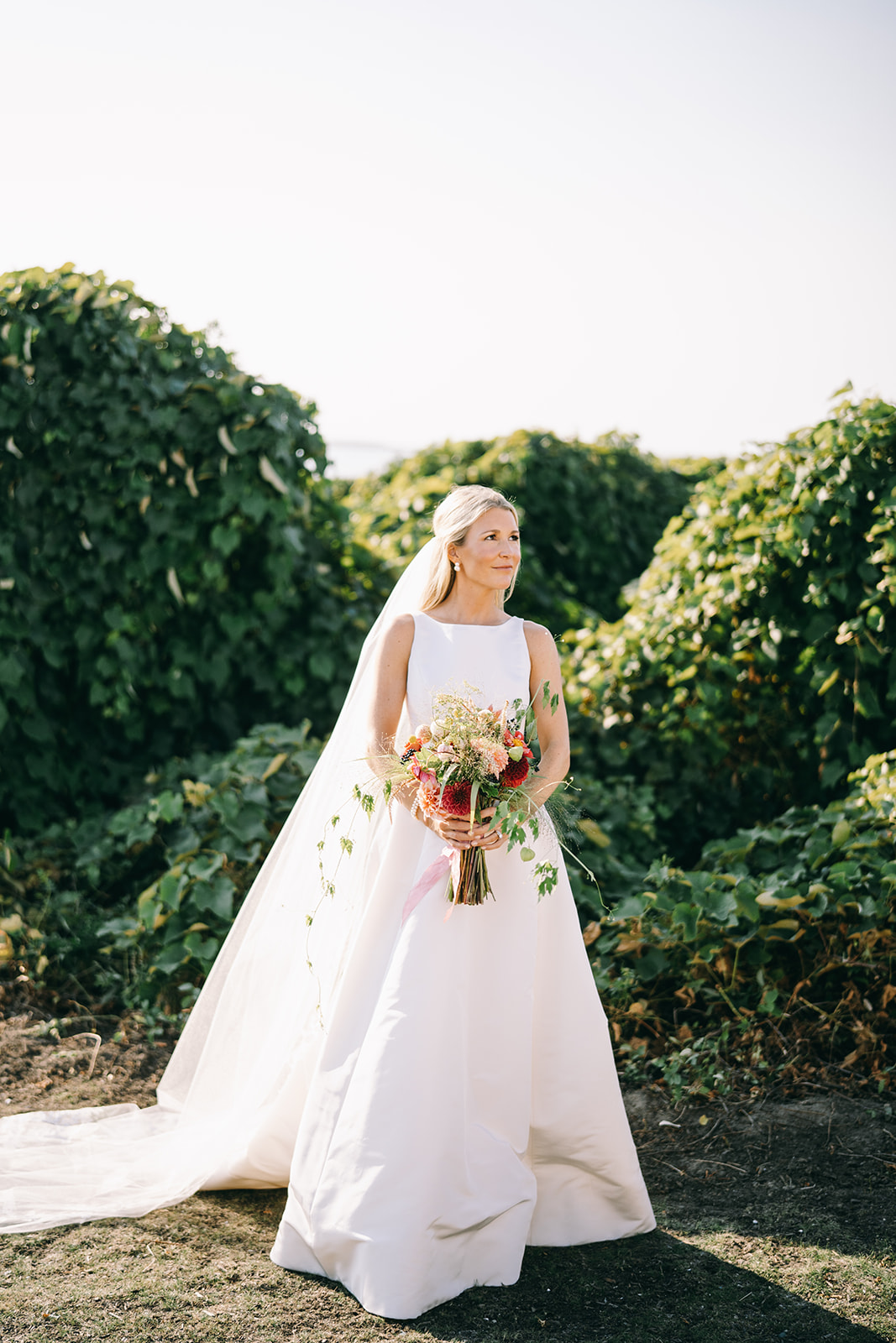 Woman in white wedding dress holding a bouquet looking off into the distance in front of greenery