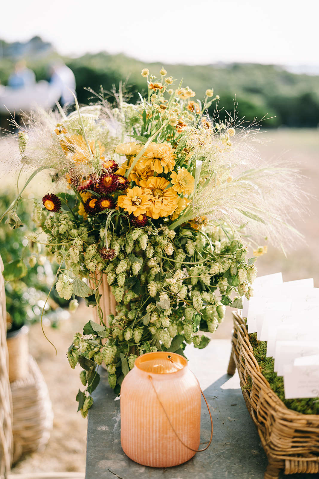 Greenery and yellow flowers in wicker basket behind an orange candle holder in 