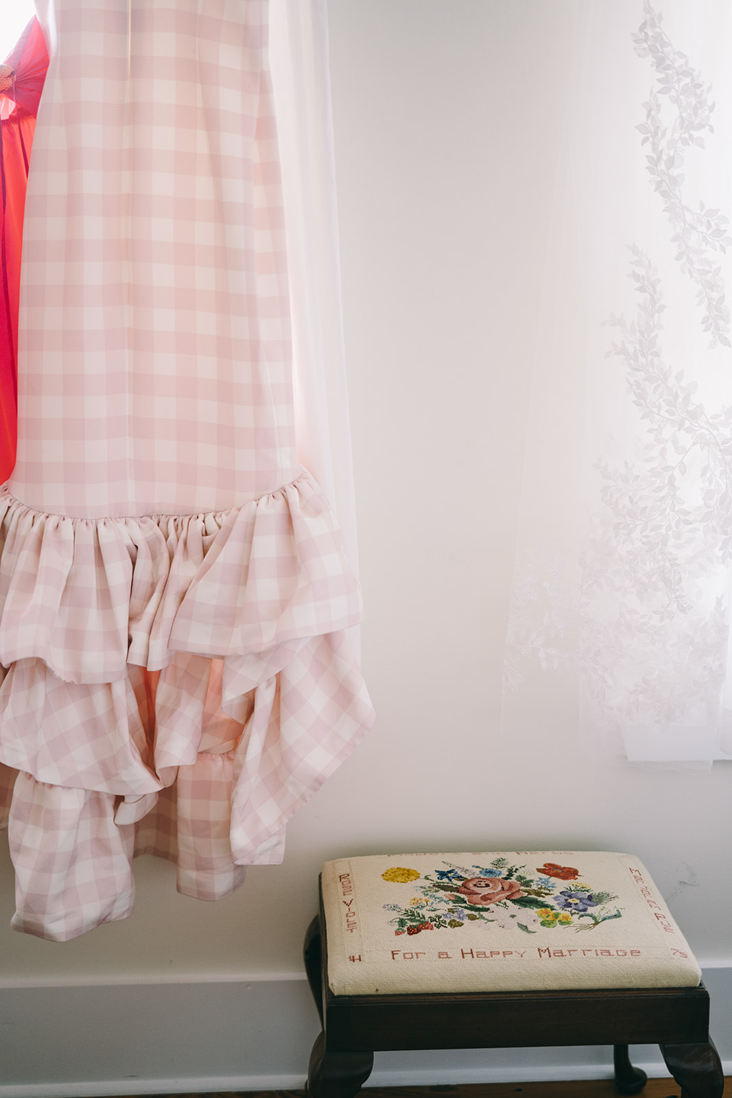 Pink and white checkered dress hanging againts white wall next to an antique stool