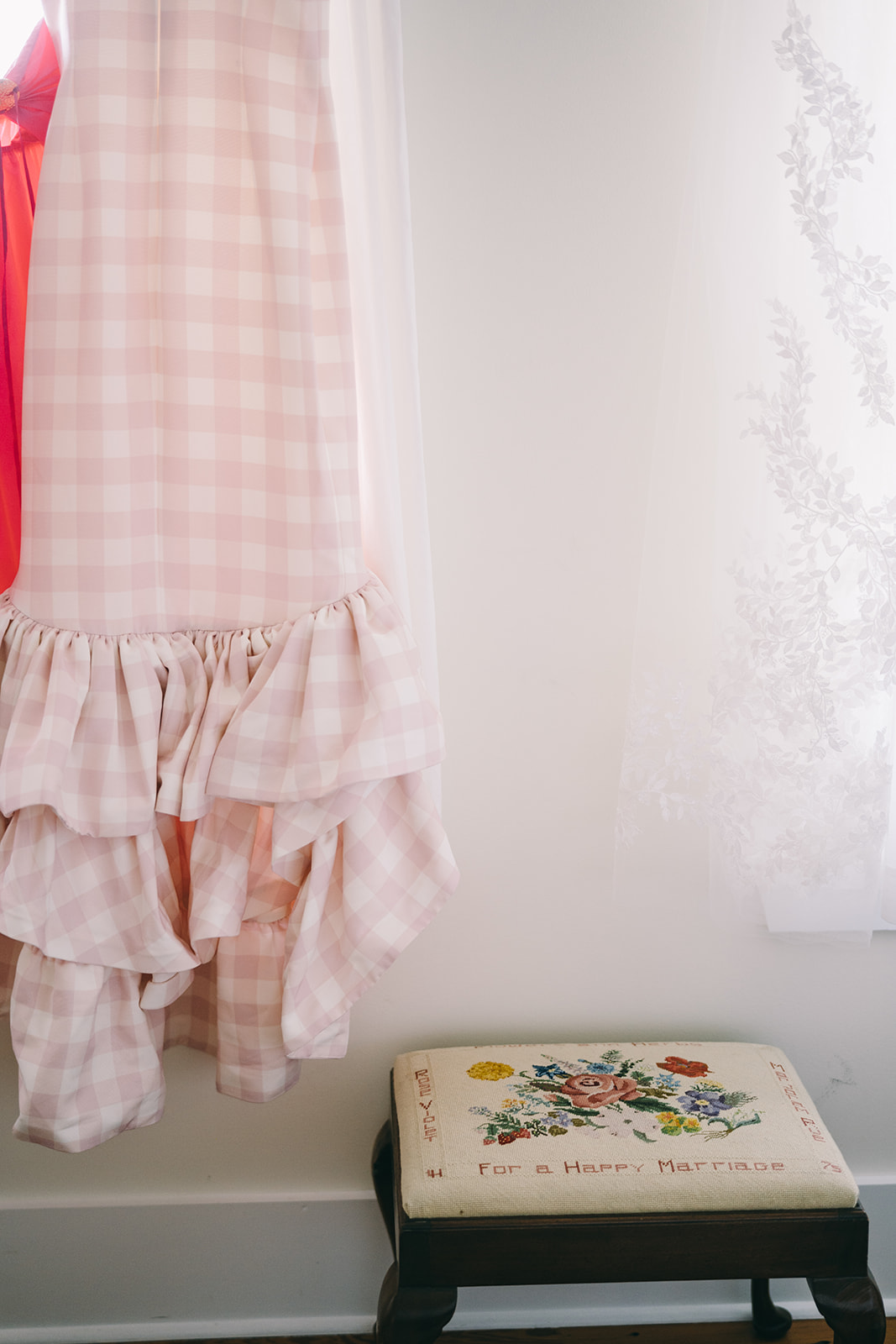 Pink and white checkered dress hanging againts white wall next to an antique stool 