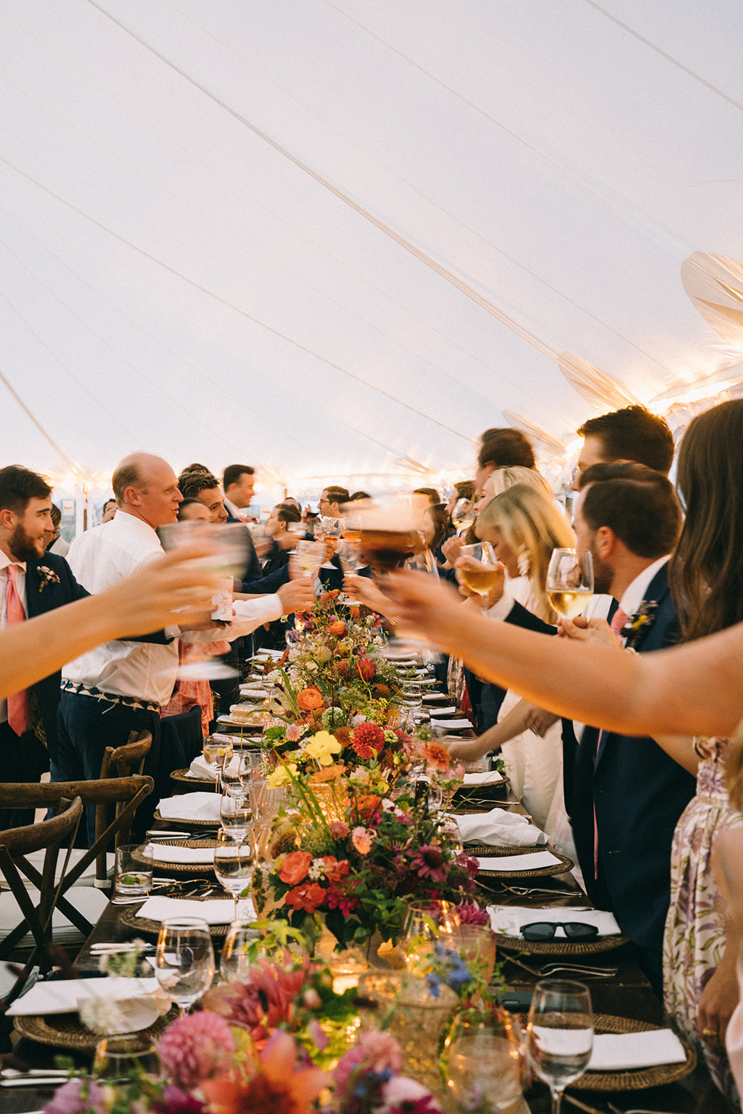 Long wedding table with colorful flowers with guests standing to make a toast 