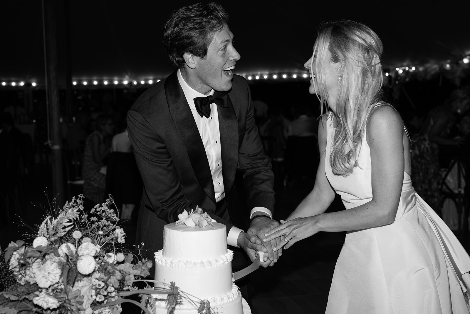 Bride and groom looking at each other laughing while cutting their cake in black and white