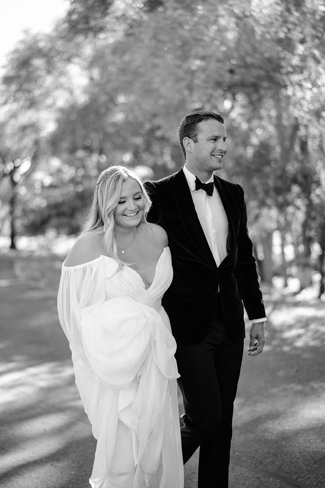 Bride and groom walking side by side, the bride is holding her dress and they are both smiling 