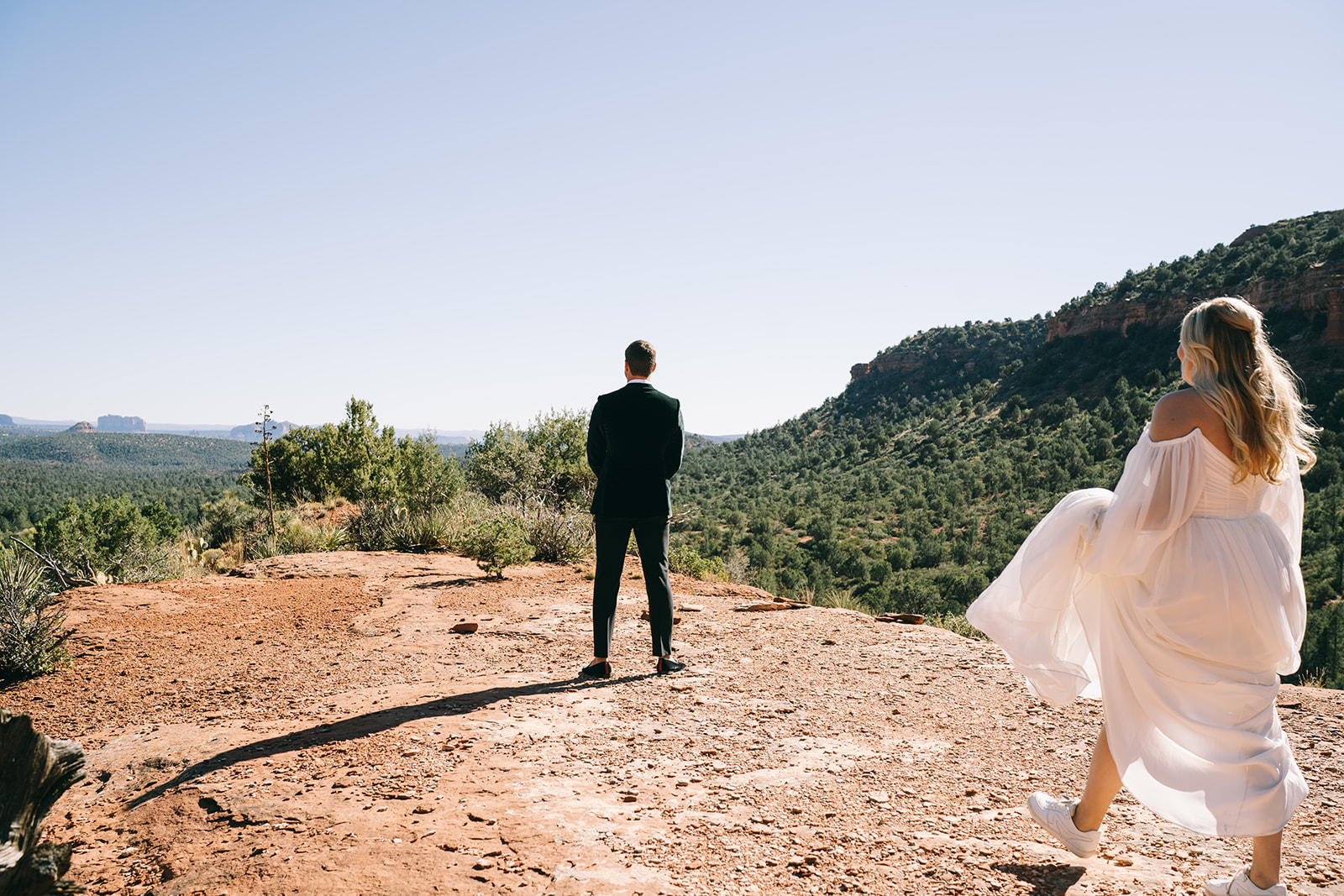 Bride holding her skirt walking towards the grom whose back is facing her surrounded by trees