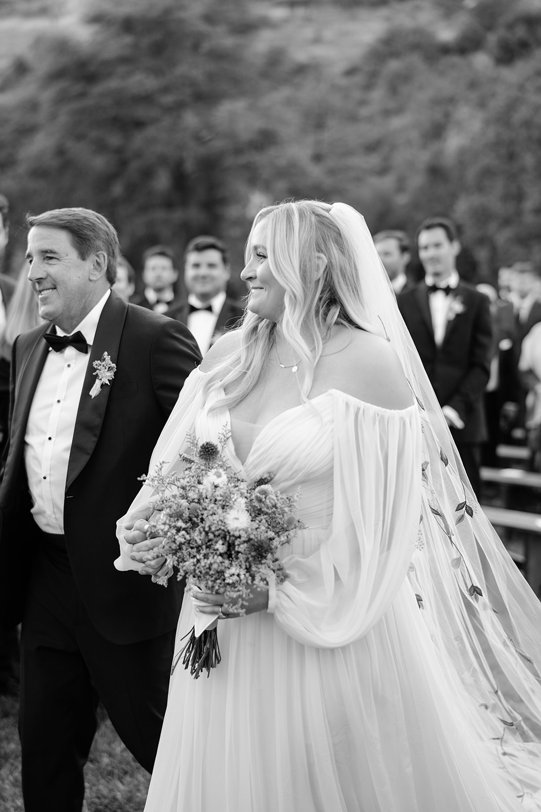 Bride holding bouquet wearing her veil walking down the aisle with her dad in black and white 