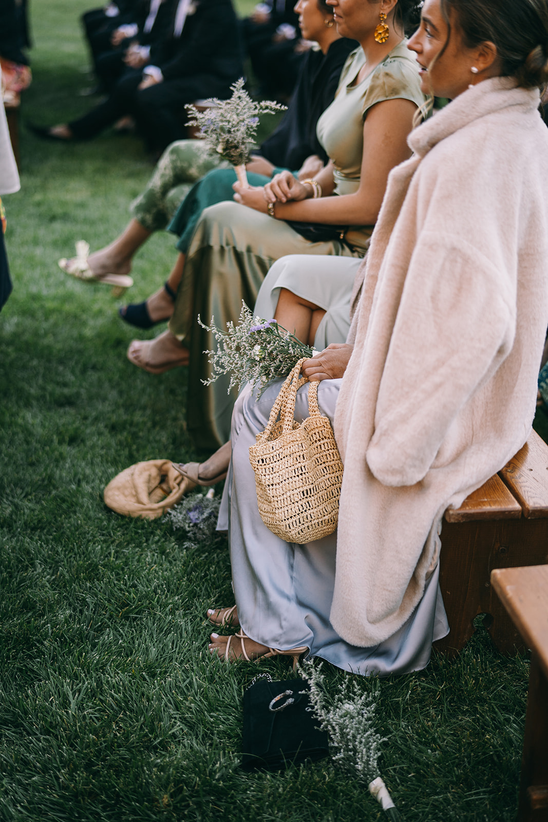 Women sitting on a bench with their legs crossed holding flowers, one has a wicker purse