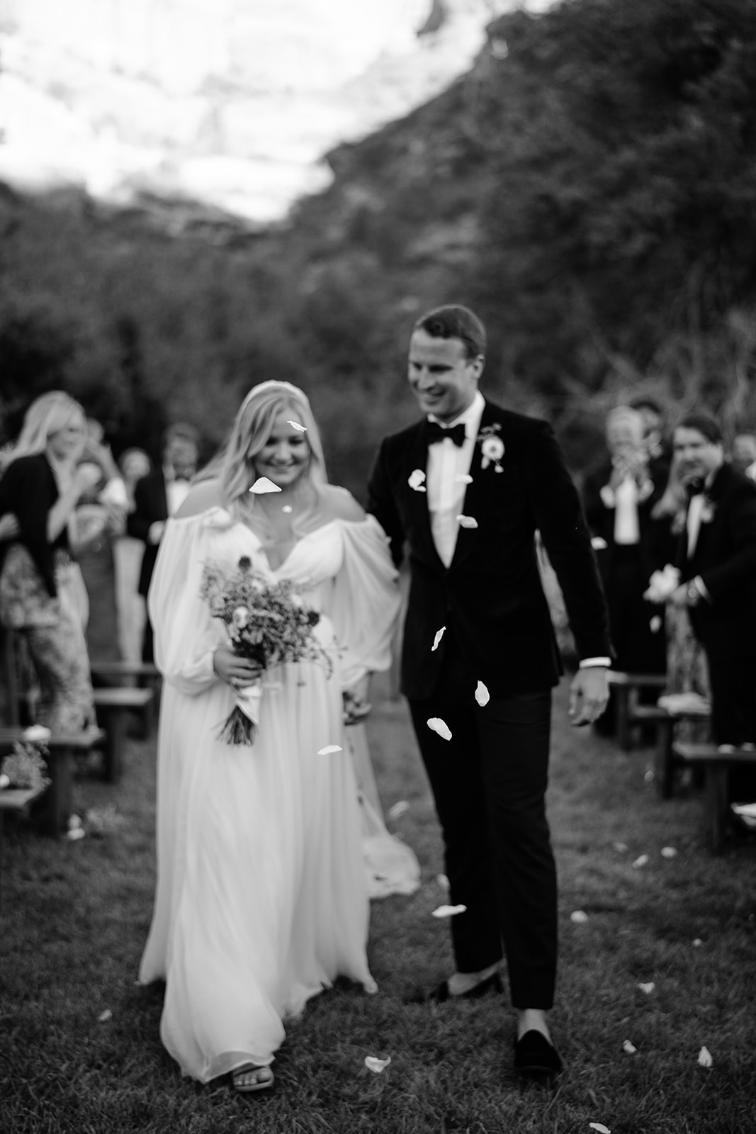 Groom looking at bride as they walk down the aisle together with flower petals being thrown at them 