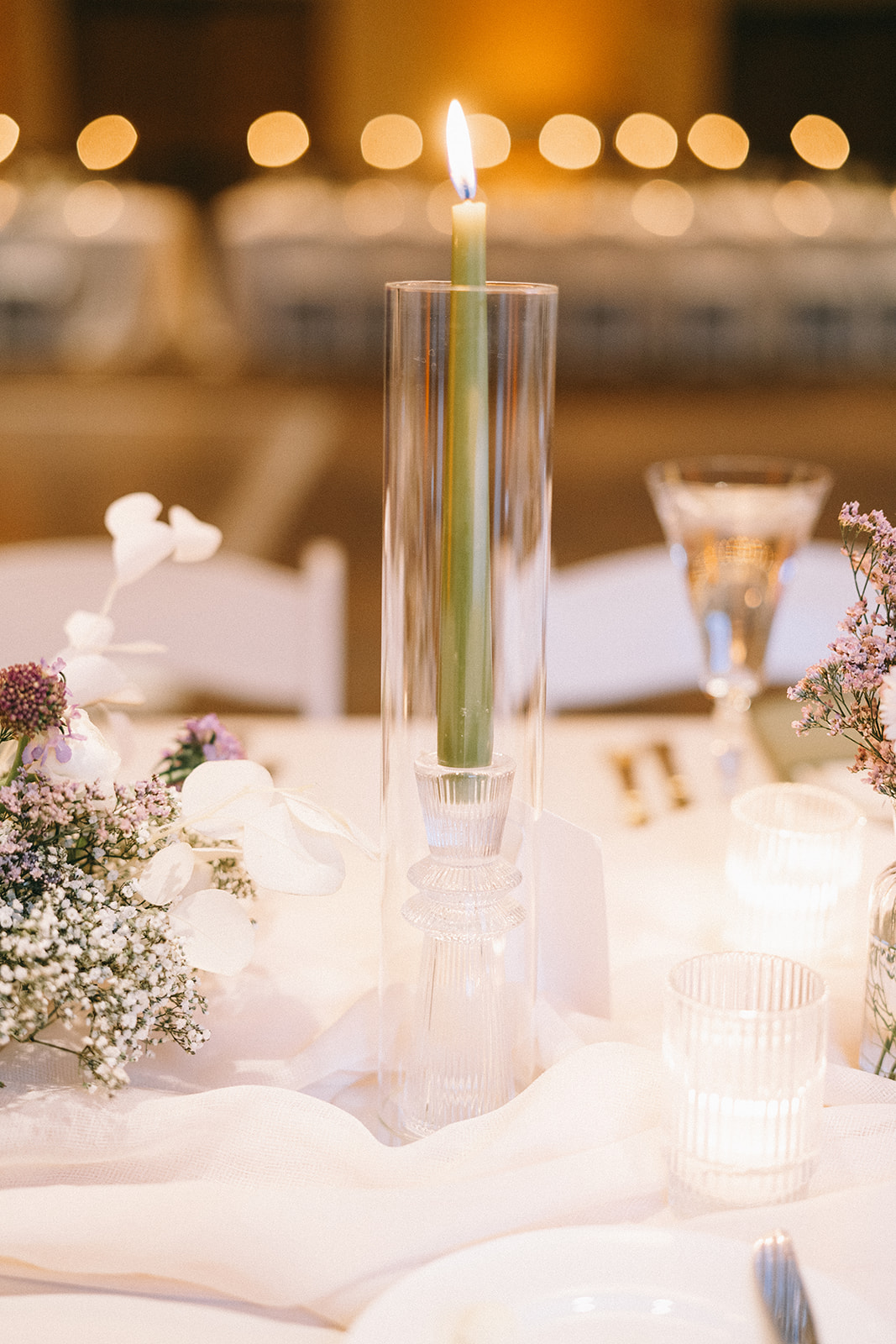 Green candle placed in a cylinder with the flame above the glass surrounded by flowers and white gauzy fabric