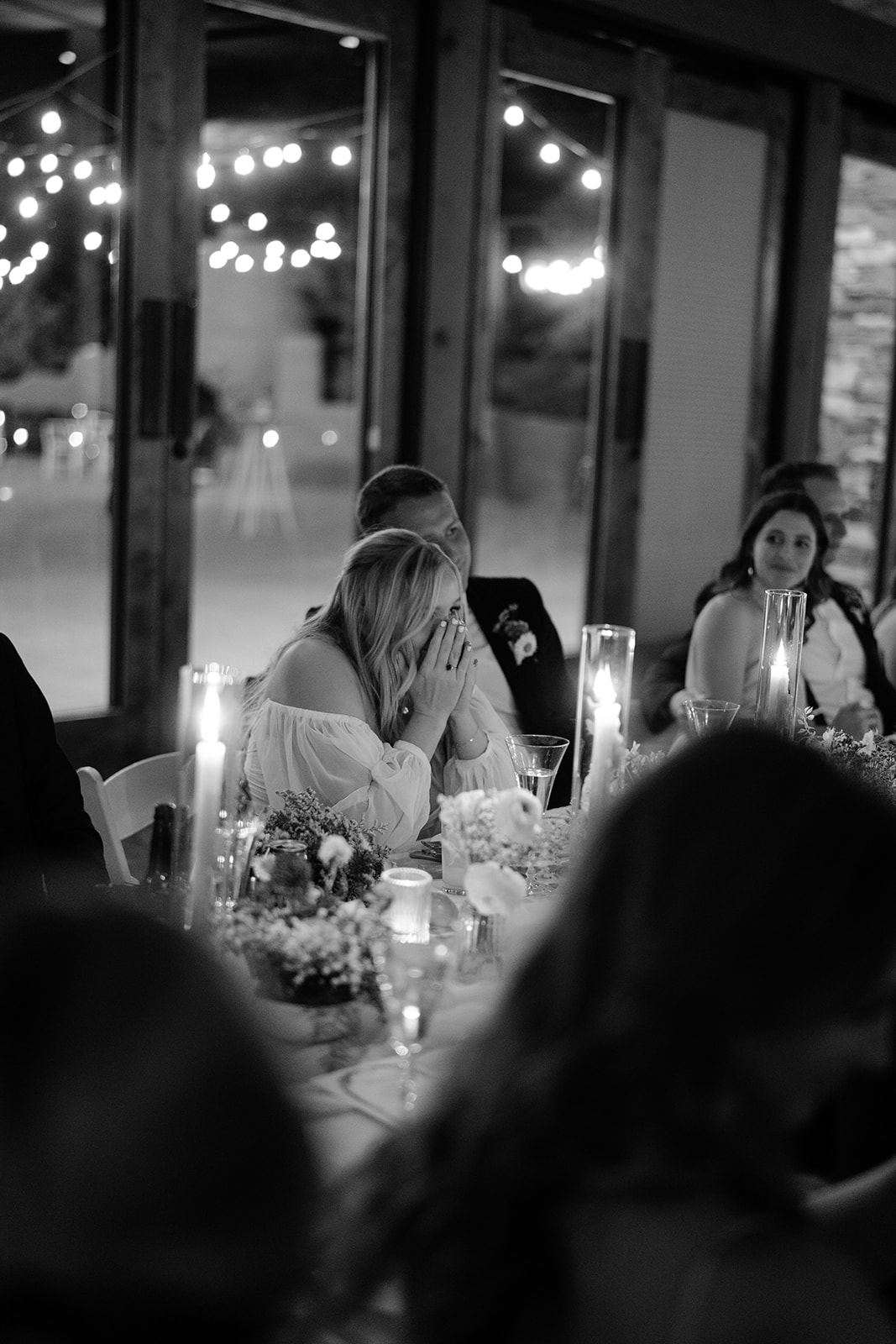 Bride covering her face laughing at table with her husband and twinkly lights in the background