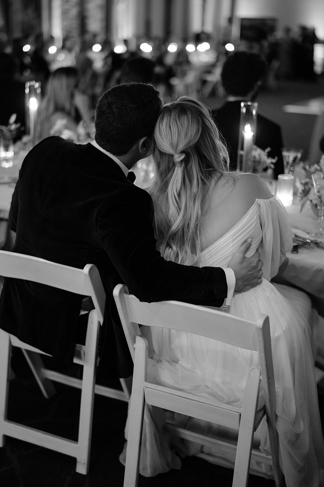 The back of bride and groom sitting together with their heads tilted in and the groom's arm around her in black amd white 