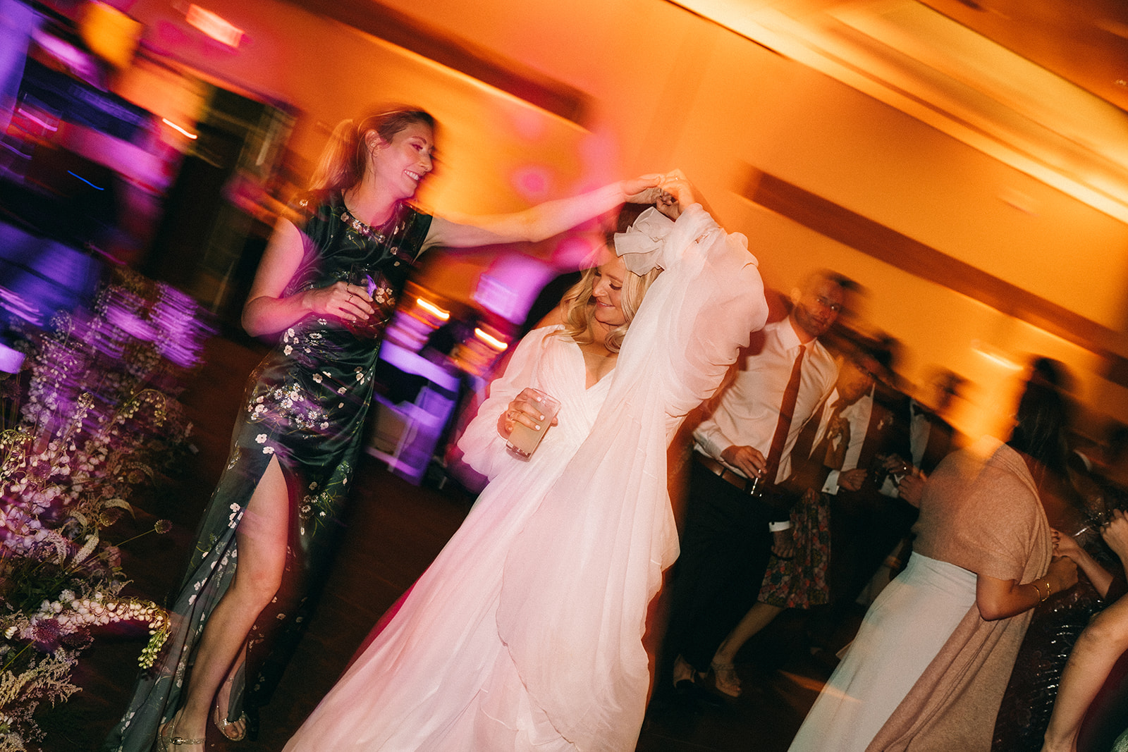 Bride being spun around by a woman in a green sparkly dress with long exposure around other guests and lights 