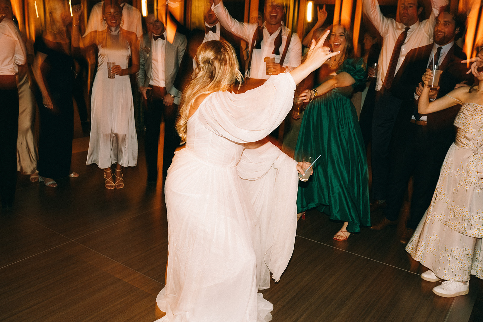 Bride in the middle of the dance floor alone making the 'rock on' hand motion while looking at her guests and laughing 