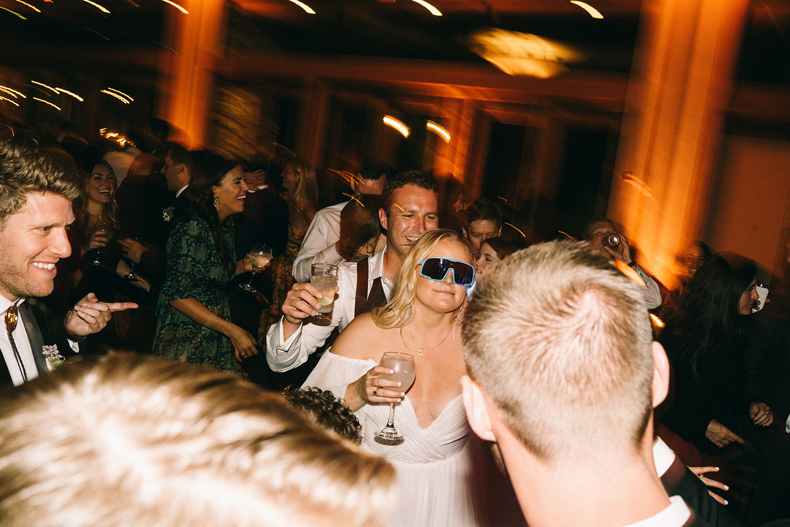 Bride in blue sunglasses holding a glass smiling and laughing with other guests while they dance 