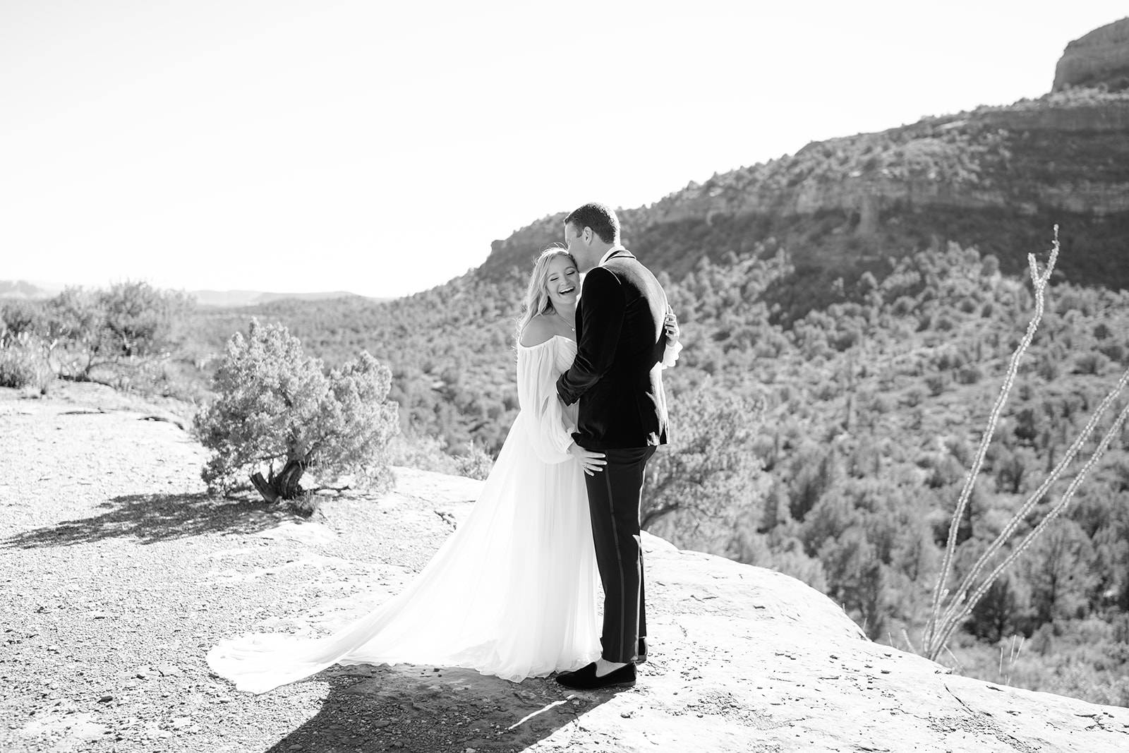 Groom kissing bride on the forehead while she is laughing with mountains and plants in background 