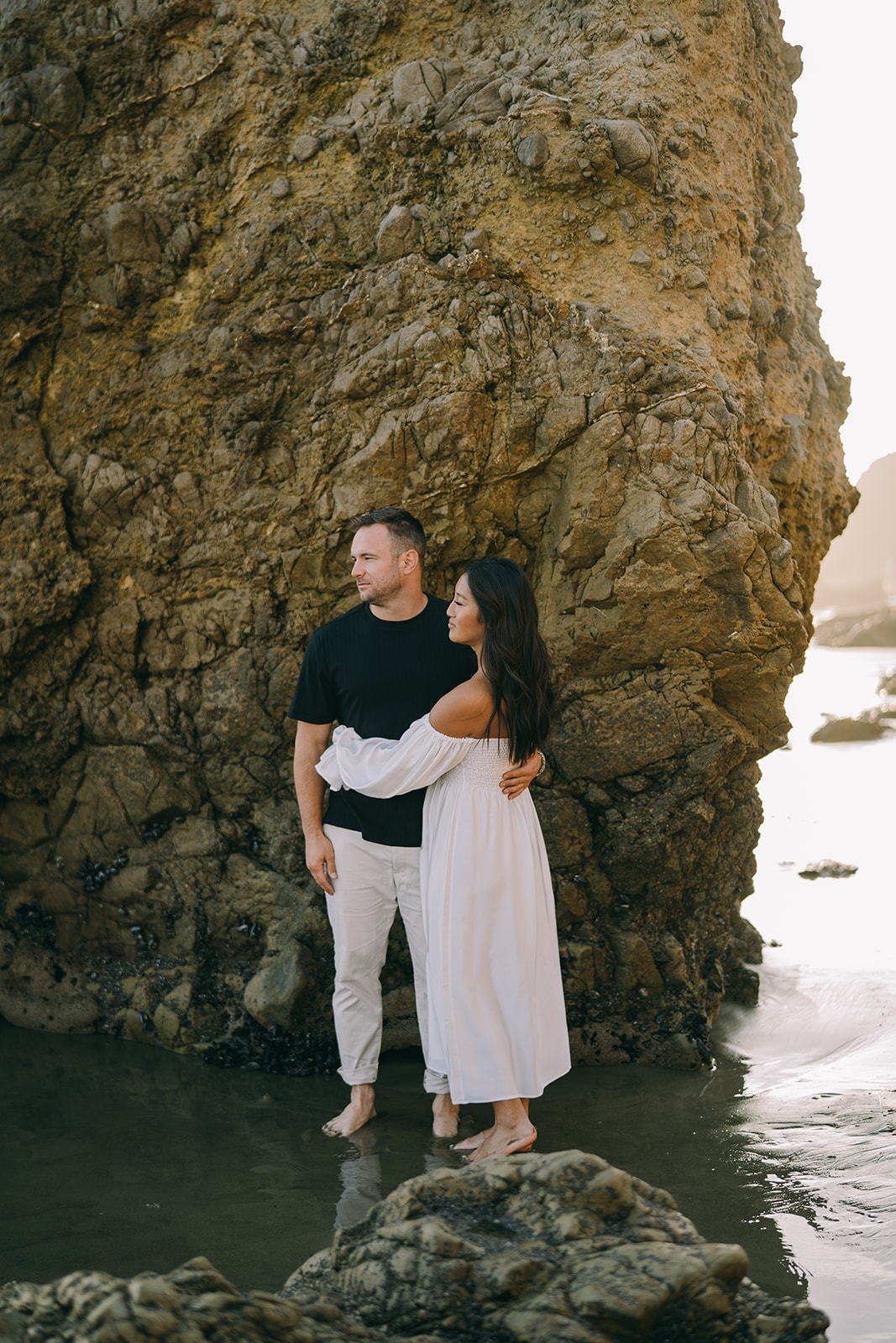 Woman in flowy dress standing with arms around a man on the coastline in Malibu