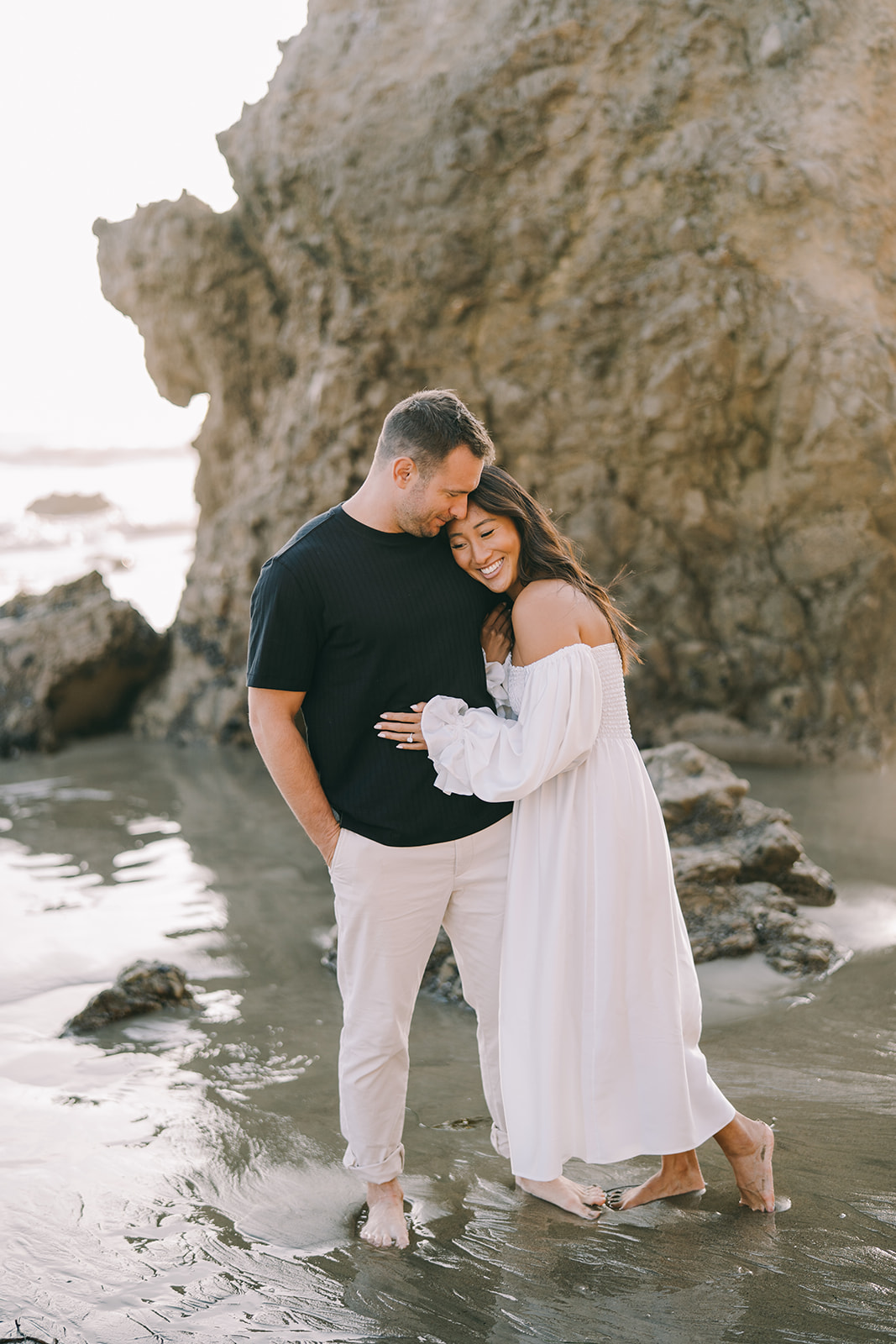 Woman in long dress leaning into man and laughing as they stand in the sand 