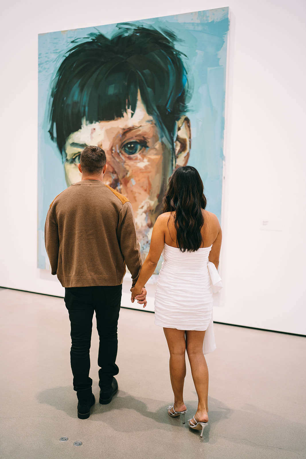 Woman in white dress and man in brown sweater looking at a piece of art of a boy with black hair