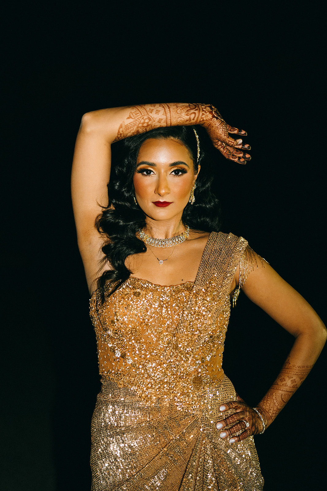 Woman with one hand over head in golden dress with hair down and henna on her hands 