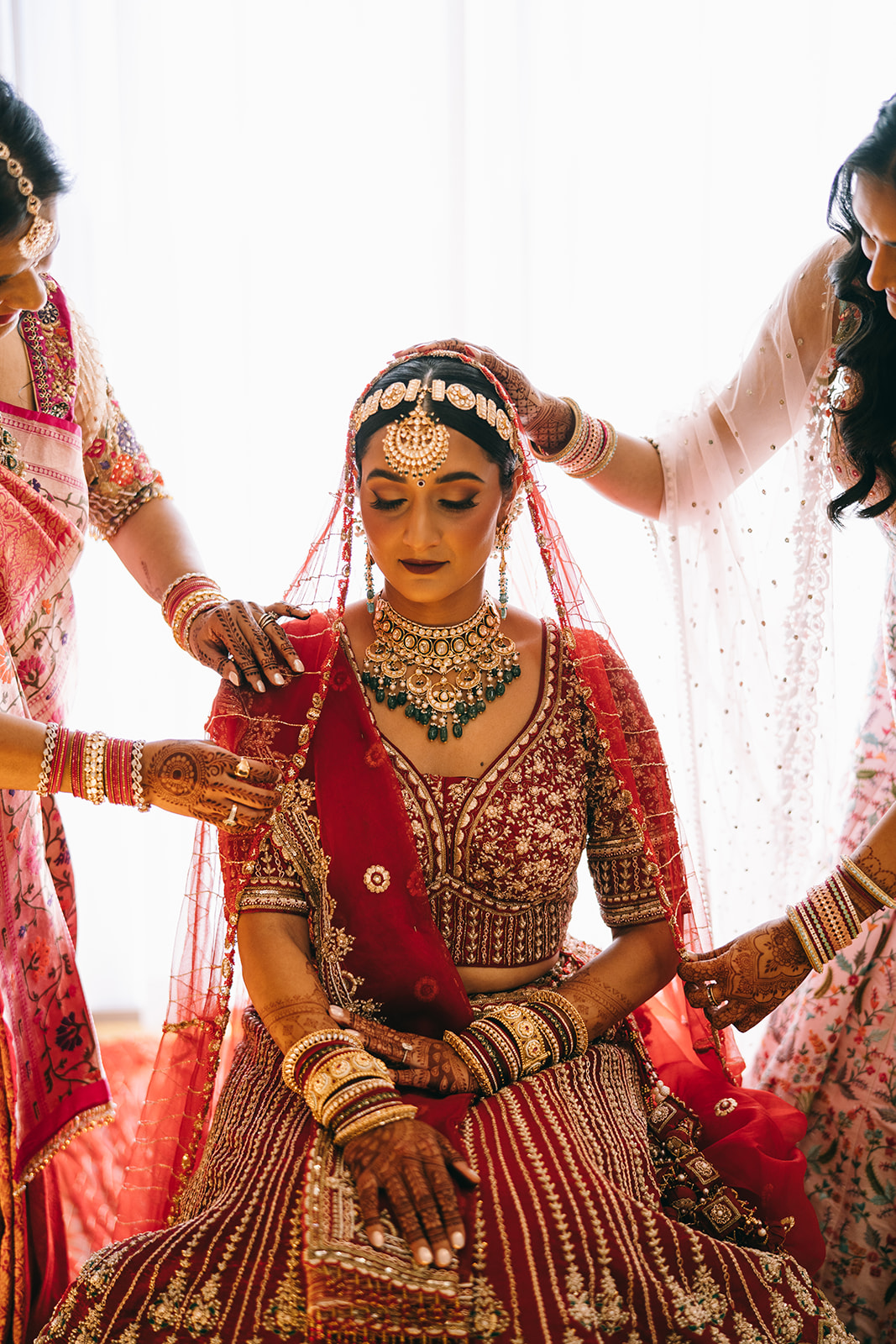 Woman in full indian wedding dress sitting down and looking down while her family helps put her veil on