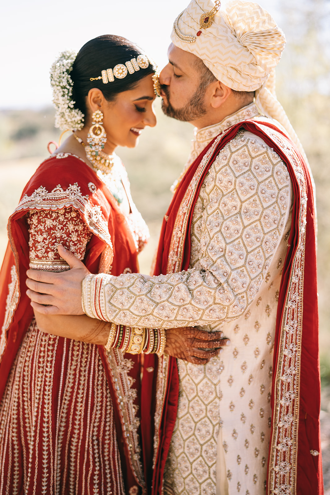 Man in golden and red outfit kissing the forhead of a woman in red and gold looking down and smiling