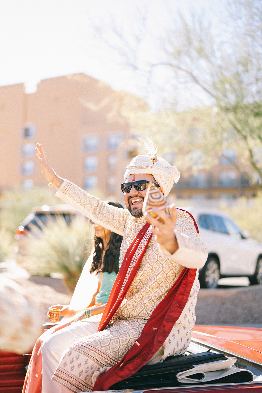 Man in gold and red outfit sitting on the back of a convertible car in sunglasses with hands outstretched smiling