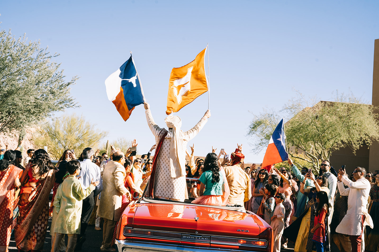 Group of people gathered around red convertible car with people waving texas state flag