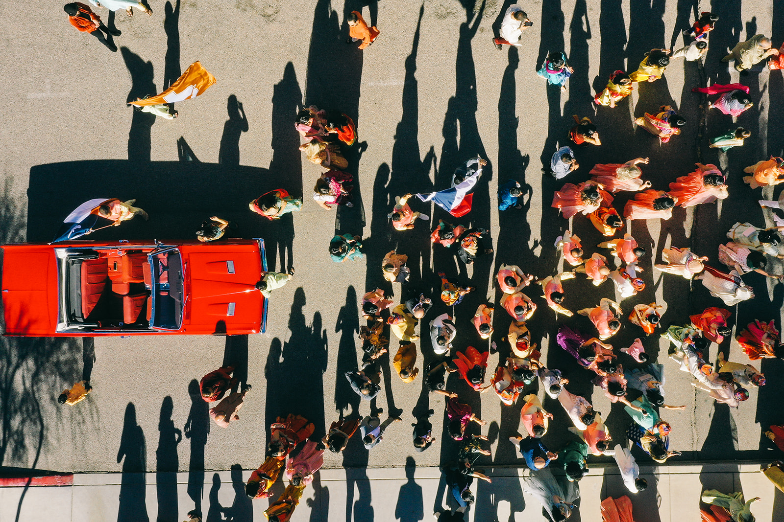 Aerial view of group of people in colorful clothing walking behind a red convertible
