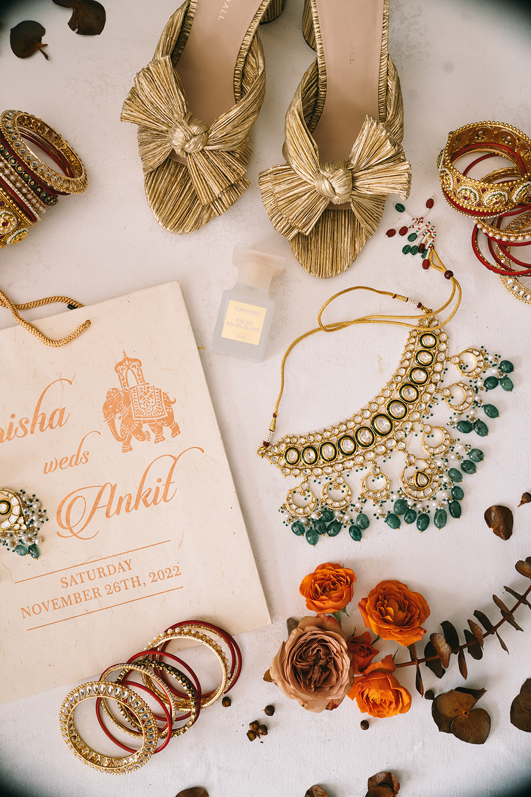 Flat lay with golden shoes, gold and green necklaces, red and gold bangles, orange roses, and a wedding invitation