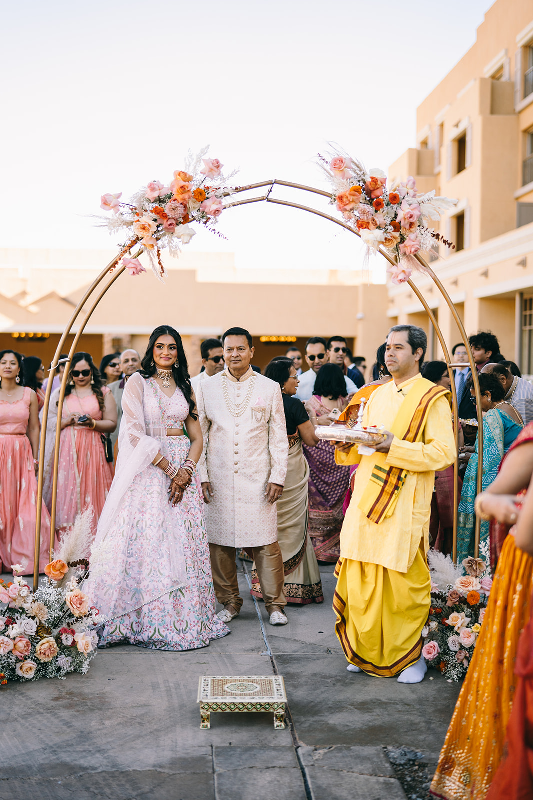 Woman in pink saari and two men underneath a understated flower arch with pink, red and orange roses