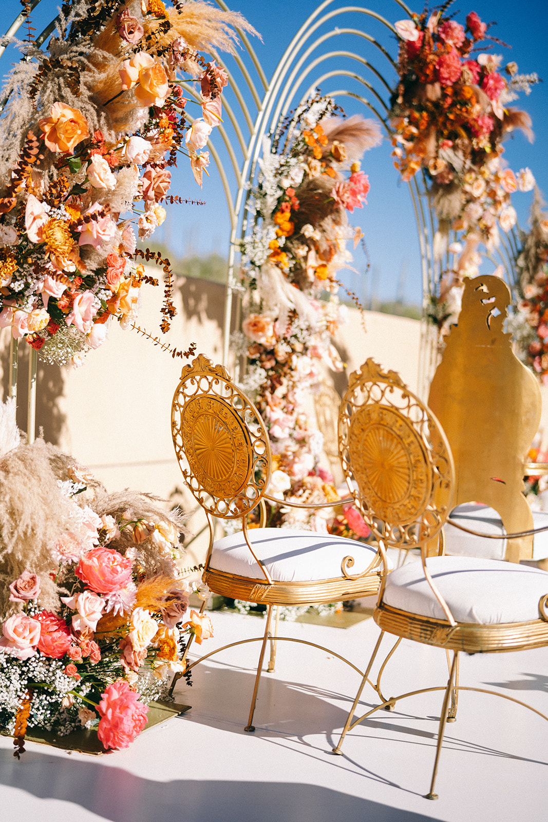Two delicate golden chairs with white seats in front of flower arches 