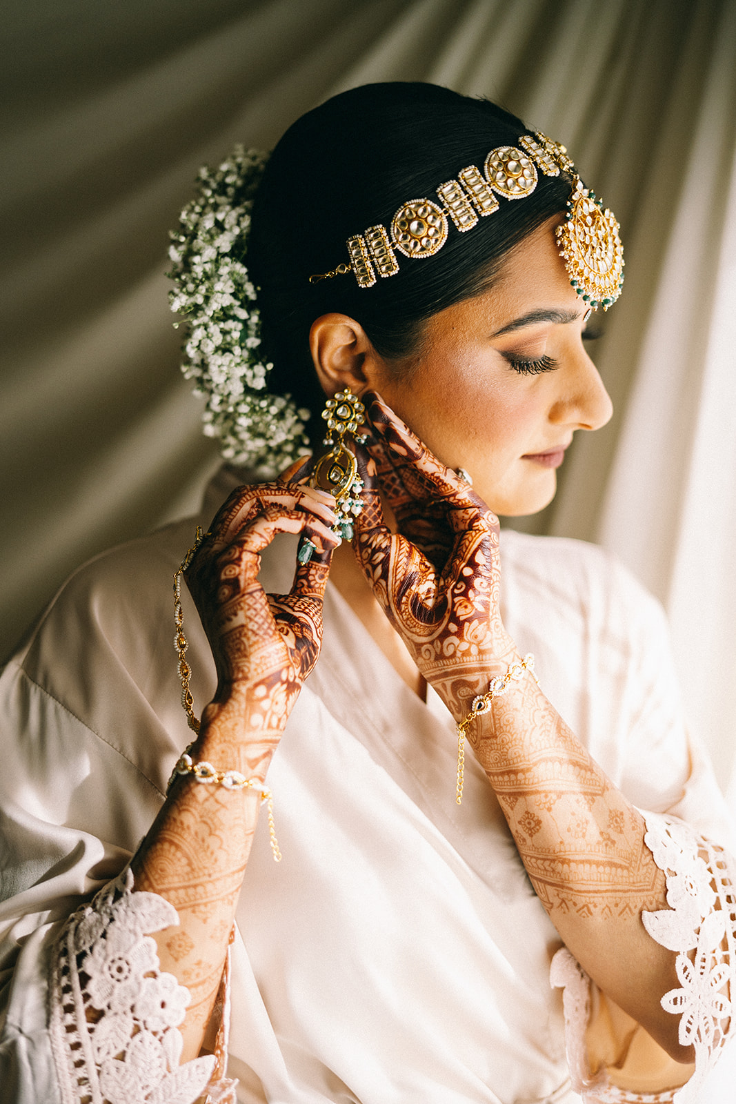 Indian woman putting on her earrings. She has henna on her hands, a golden headress, and little white and green flowers in her hair that is pulled up