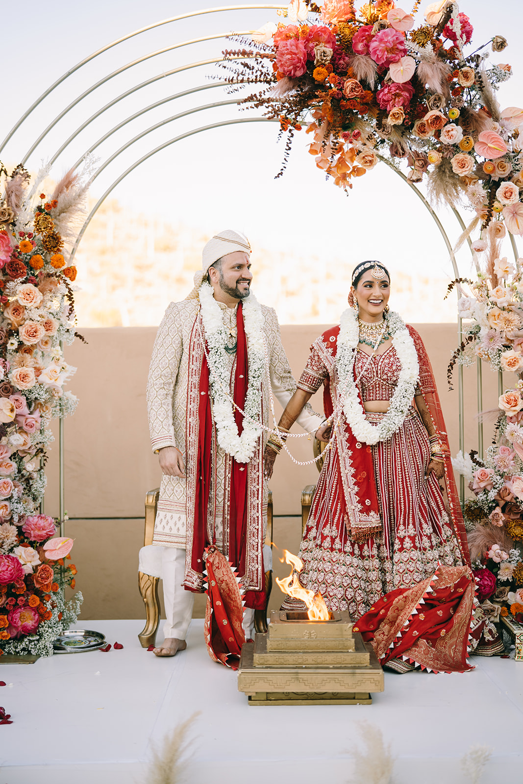 Indian bride and groom with flower necklaces standing under flower arch smiling while tiny flam in golden box burns 