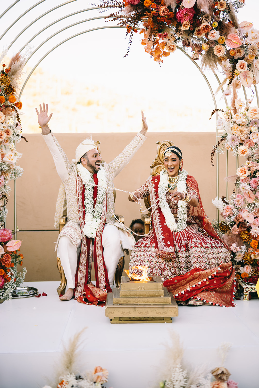 Indian groom with his hands up and cheering and indian bride is smiling with strings of flowers in her hands