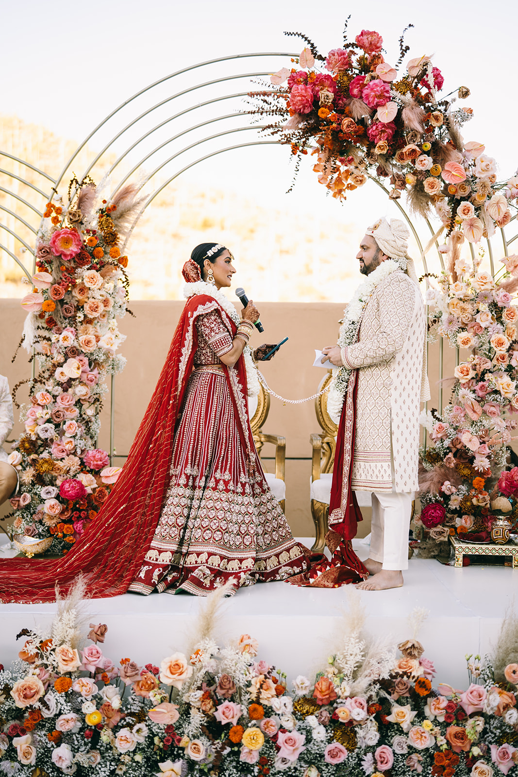 Indian bride holding mircophone and looking at her groom under flower arch
