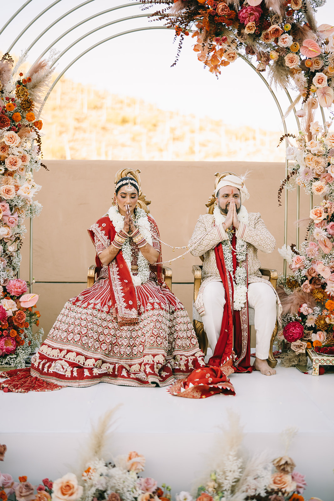 Indian bride and groom sitting next to each other with hands folded in prayer in front of their faces 