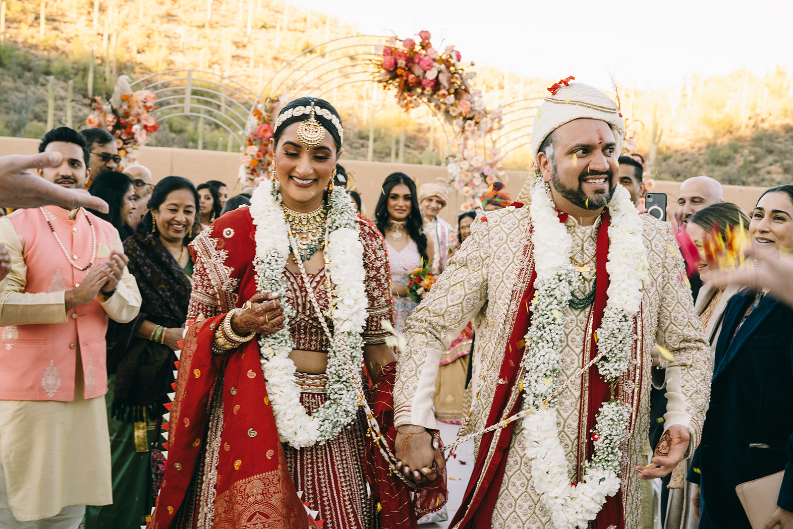 Indian couple at their wedding with flower necklaces holding hands and walking in a procesion while people throw yellow flower petals