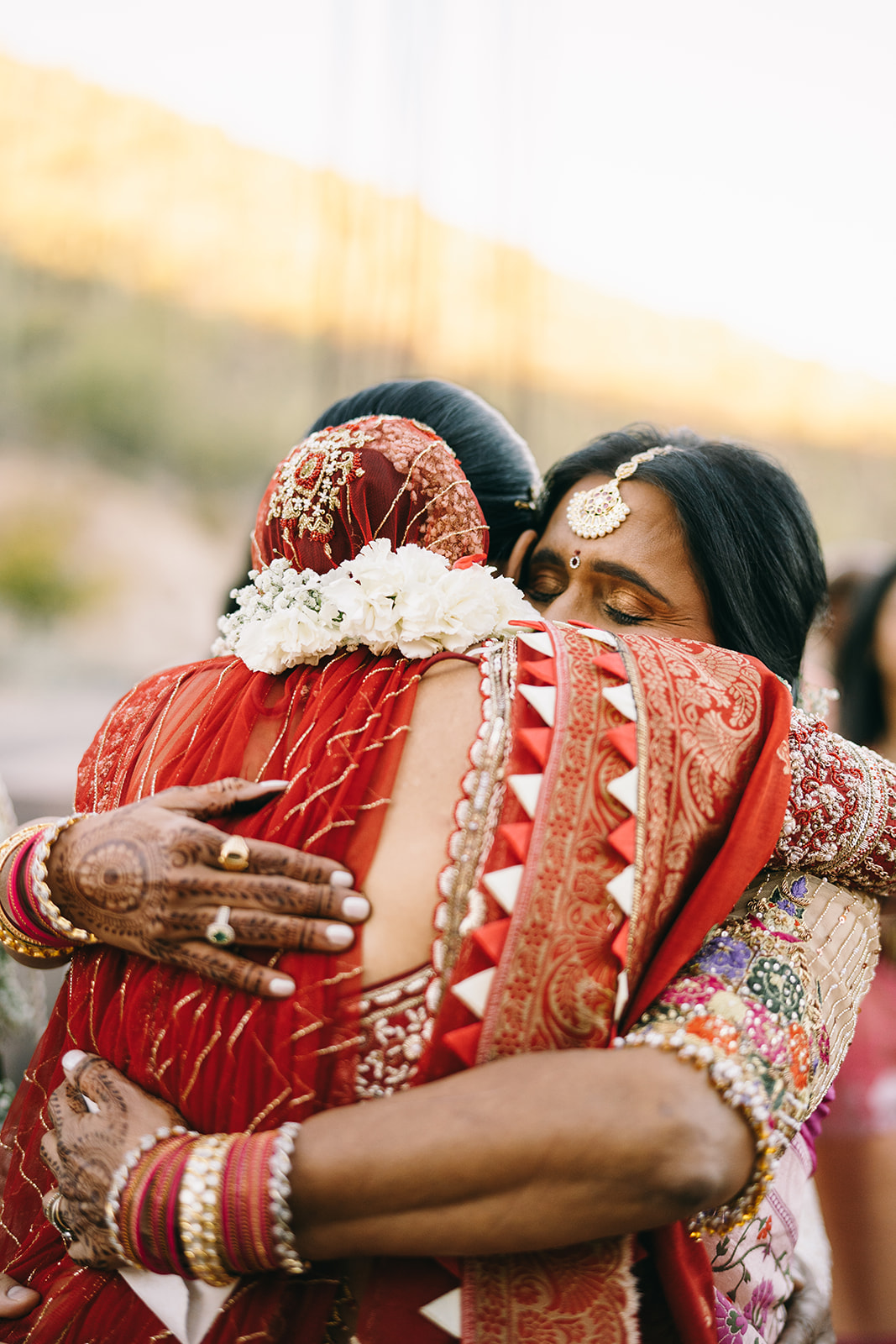 Indian bride being embraced by an older Indian woman with red and gold bangles and henna on her hand