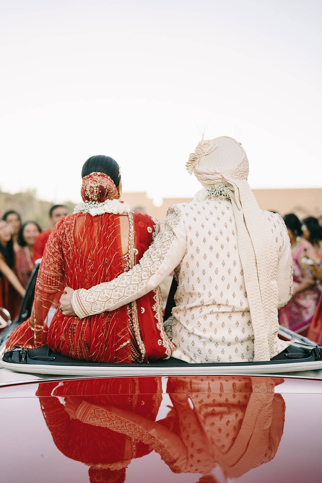 View of indian bride and groom sitting on red convertible from the back and the groom has his arm around the bride 