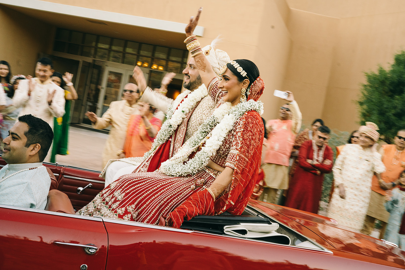 Indian bride and groom smiling and waving from the back of a convertible as they are being driven 